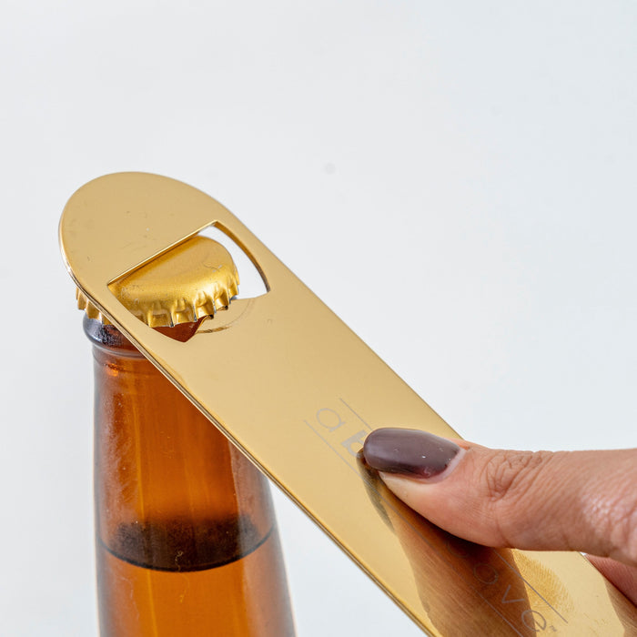 A gold bar blade bottle opener held by a hand, opening a brown glass bottle.