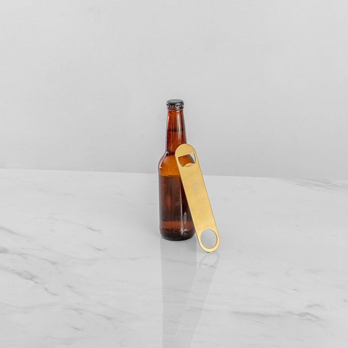 Gold bar blade bottle opener leaning against a brown beer bottle on a white marble surface.