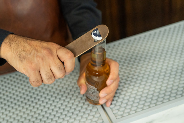 Brushed bronze-toned stainless steel bar blade bottle opener prying the cap off a beer bottle.