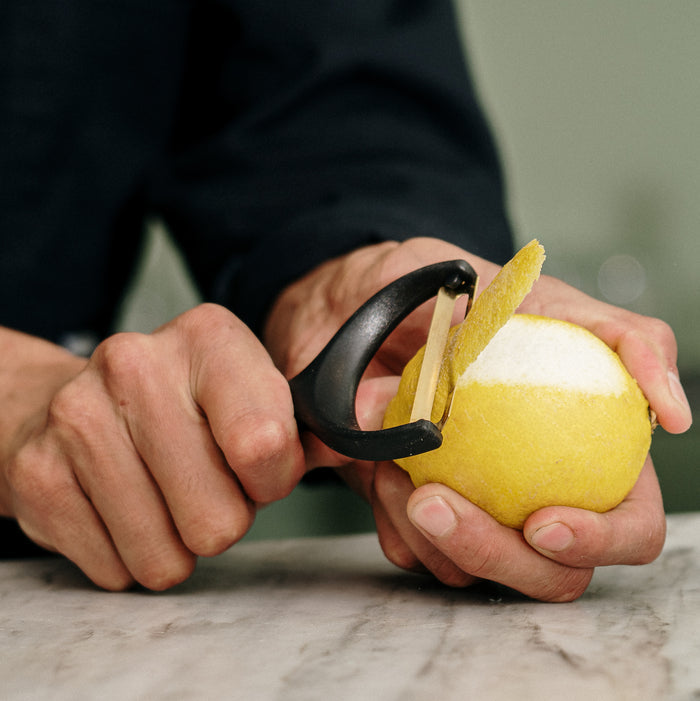 Black-handled gold citrus peeler held by hands, actively peeling a lemon over a marble surface.