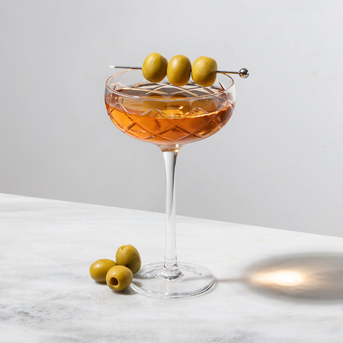 A clear diamond-cut coupe glass filled with an amber cocktail, garnished with three green olives.