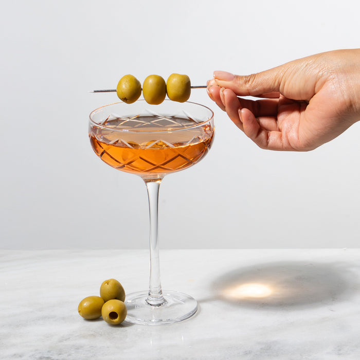 Clear diamond-cut coupe glass filled with amber liquid, a hand garnishing with three green olives.
