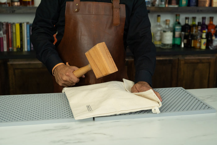 A bartender in a leather apron holds a wooden mallet above a natural canvas Lewis bag.